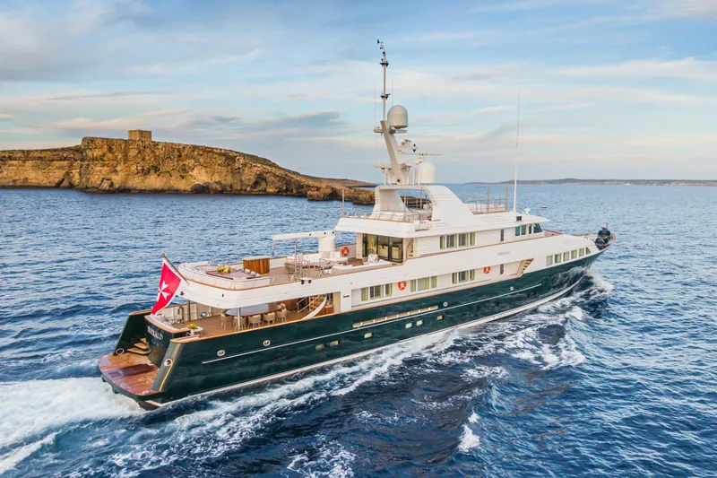Emerald Yacht Photos Pics Luxurious 1990 Feadship yacht cruising near a rocky coastline under a clear sky.