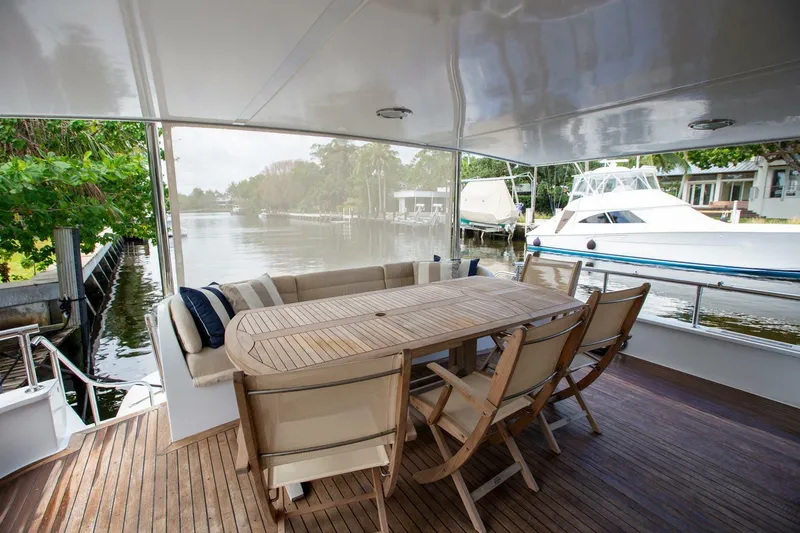 Painkiller Yacht Photos Pics Outdoor dining area on 1999 Tarrab Motor Yacht with wooden table and chairs, docked by a serene river.