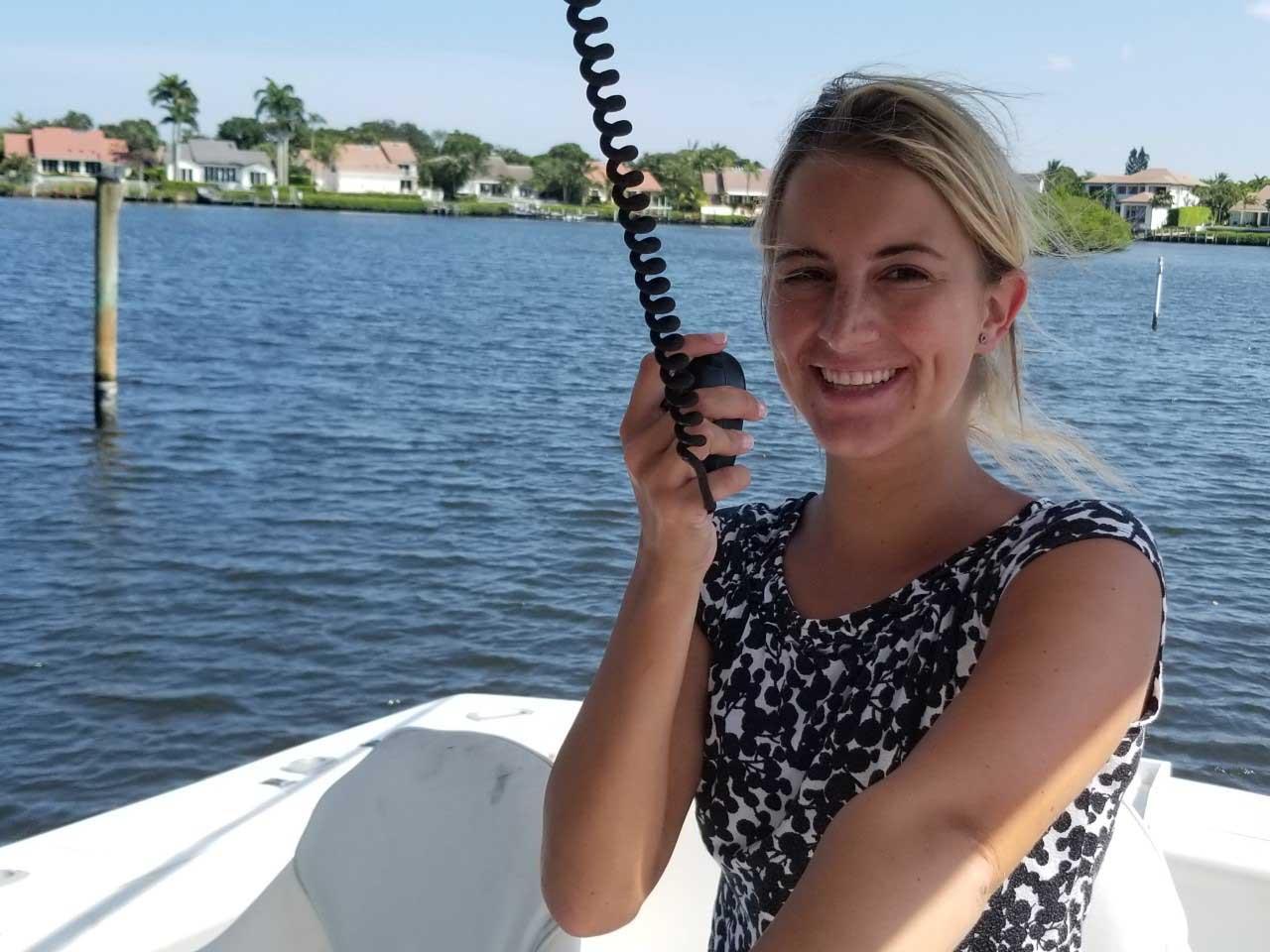 A woman smiling on a boat, holding a radio, with waterfront homes in the background.