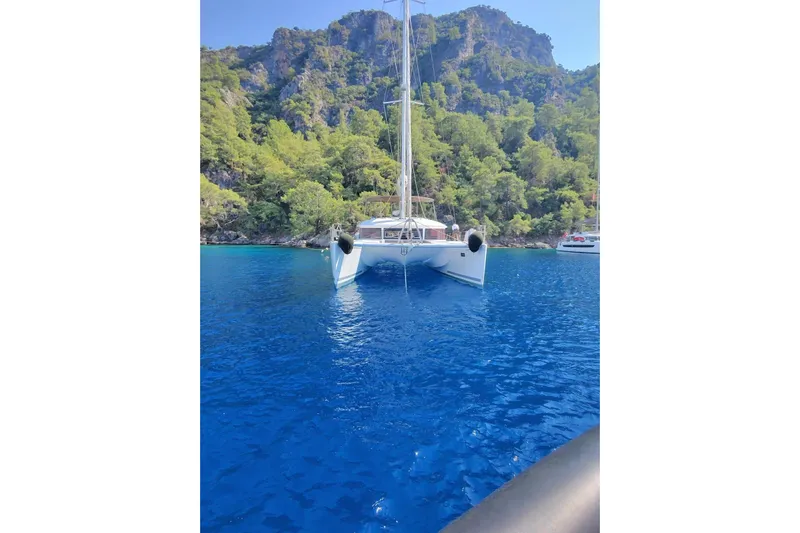 Yacht Photos Pics Lagoon 560 S2 catamaran sailing in clear blue waters near a lush, rocky coastline.