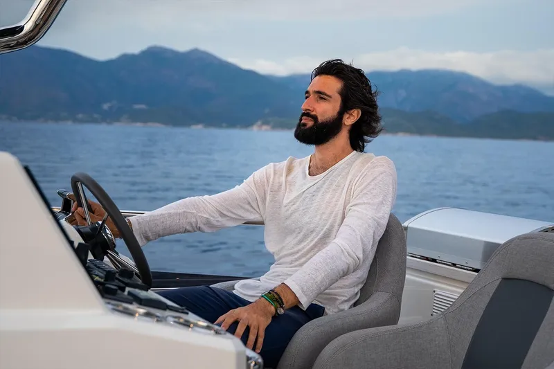  Yacht Photos Pics Man steering 2026 Beneteau Grand Trawler 63 on open water, with scenic mountain backdrop.
