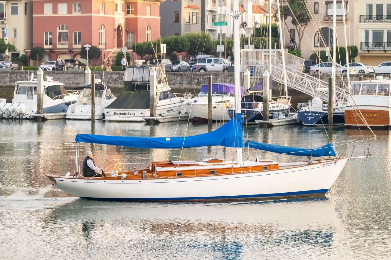 1938 Philip Rhodes Cutter sailboat with blue cover in a marina, surrounded by other boats.