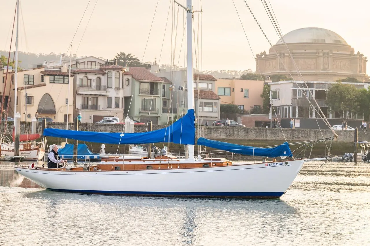 Vintage 1938 Philip Rhodes Cutter sailboat with blue sails in a scenic harbor setting.