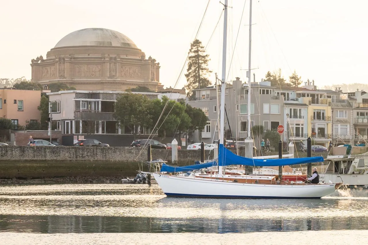 Vintage 1938 Philip Rhodes Cutter sailboat on calm waters near historic architecture.