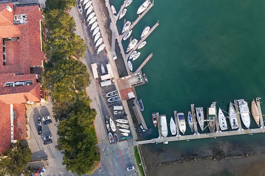 Aerial view of marina with boats docked, adjacent to red-roofed buildings and parking area.