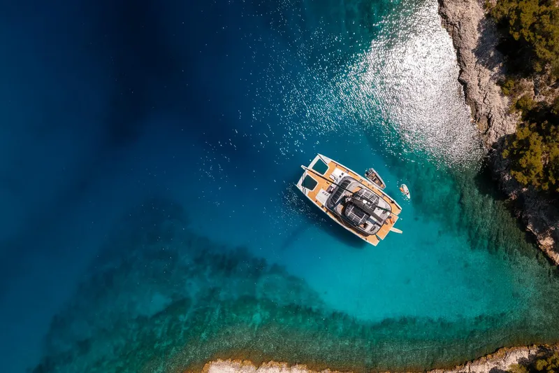 N+1 Yacht Photos Pics Aerial view of 2024 Sunreef 70 Eco yacht in clear turquoise waters near rocky coastline.