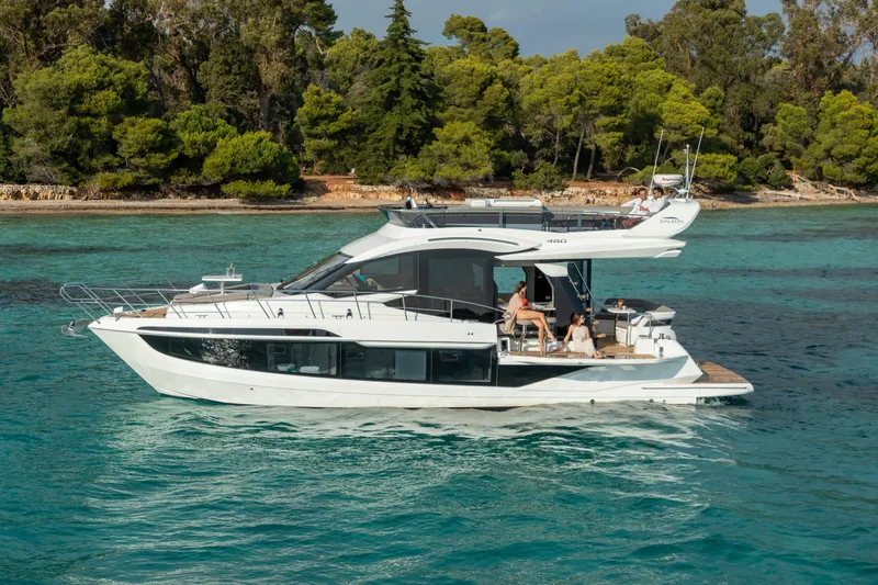  Yacht Photos Pics 2026 Galeon 480 Fly yacht cruising in turquoise waters near a lush, forested shoreline.