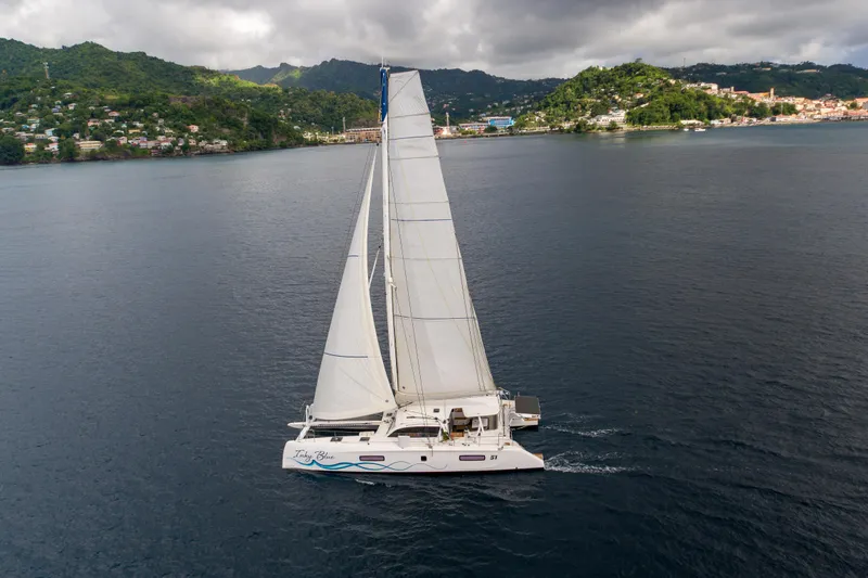 Inky Blue Yacht Photos Pics 2020 Outremer 51 catamaran sailing near lush, hilly coastline under cloudy skies.