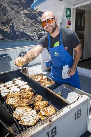 Truth Yacht Photos Pics Man grilling food on a boat, wearing sunglasses and apron, with scenic background.