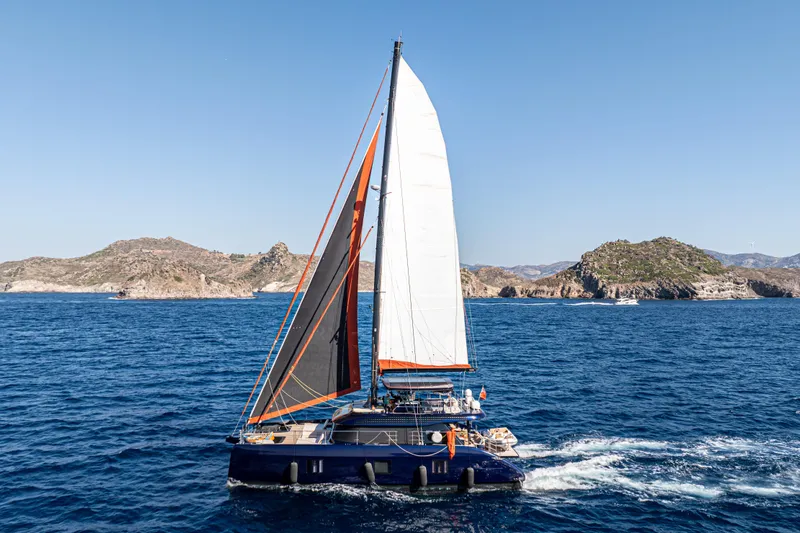  Yacht Photos Pics 2024 Sunreef 60 Eco catamaran sailing in clear blue waters near rocky coastline.