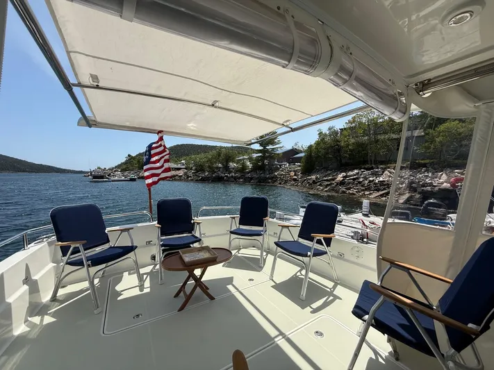 Wahoowa Yacht Photos Pics 2019 Back Cove Downeast boat deck with chairs, table, and American flag, near rocky shoreline.
