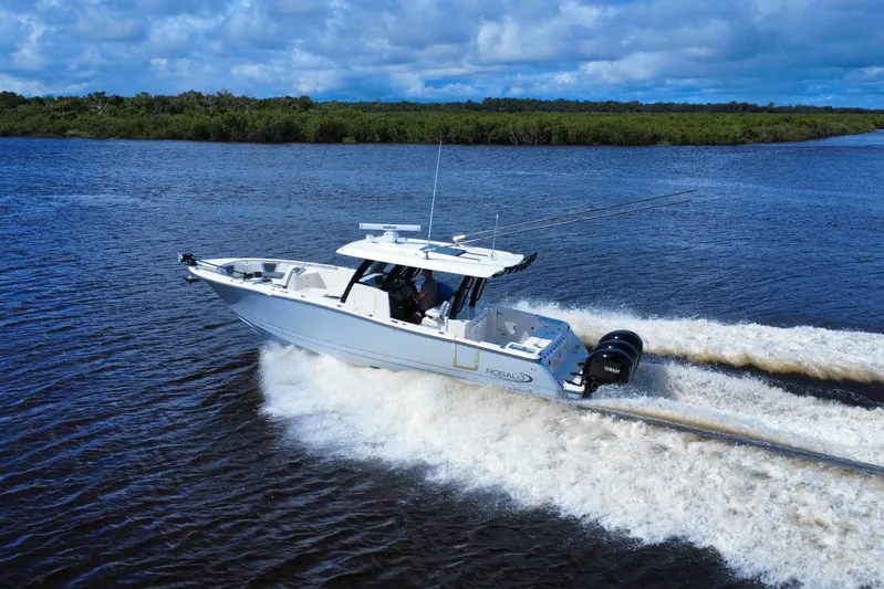 No Joke Yacht Photos Pics 2022 Robalo R360 Center Console boat cruising on a river under a cloudy sky.