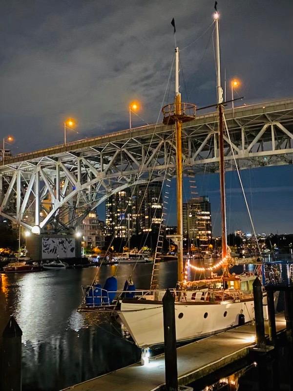 Staysail schooner docked at night under a lit bridge, city skyline in background.