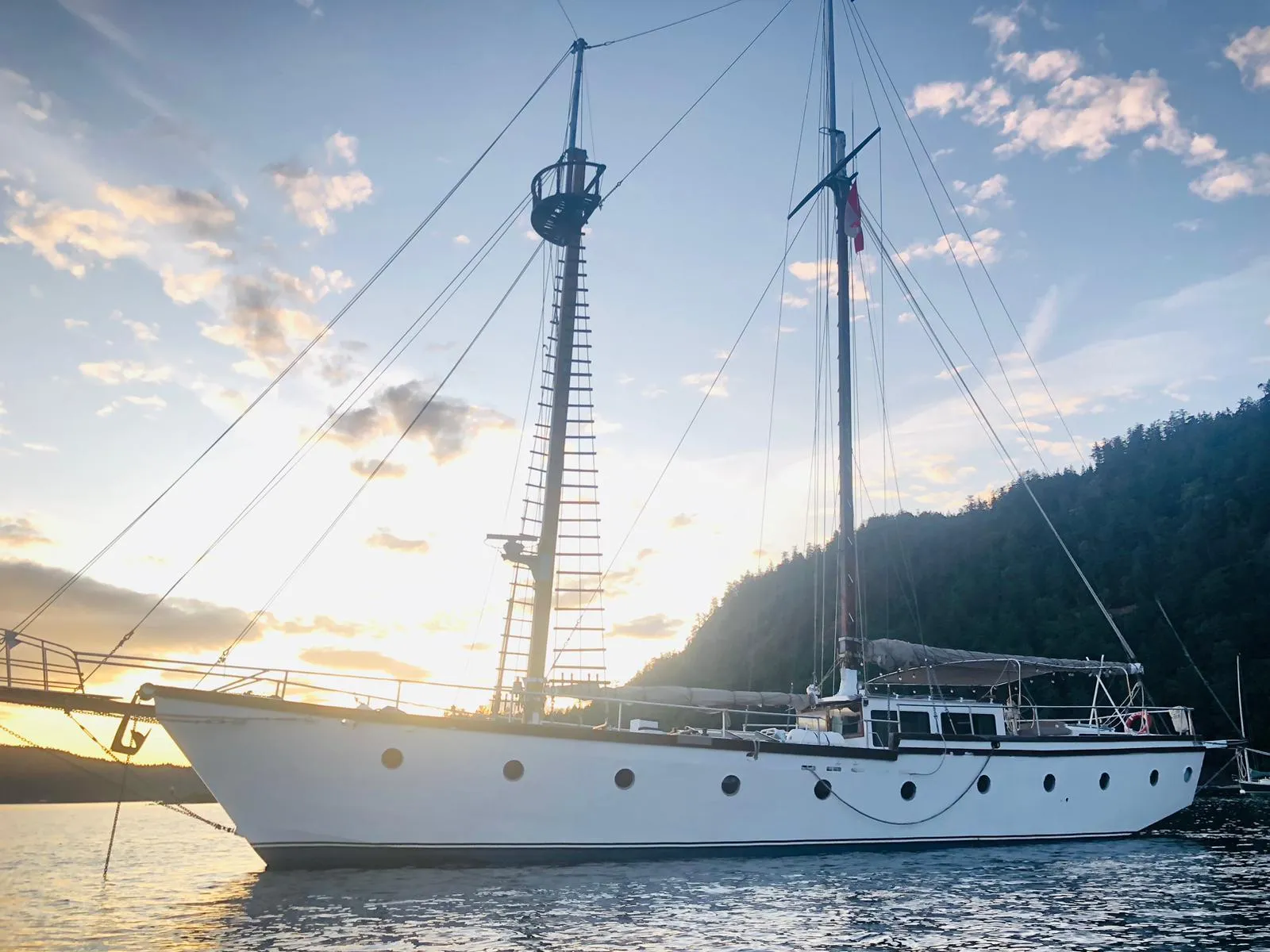 1975 Custom Staysail Schooner at sunset, anchored near a forested coastline.