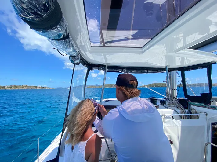 Good Times And Tan Lines Yacht Photos Pics Couple enjoying a sunny day on a 2023 Lagoon 42 catamaran, sailing in clear blue waters.