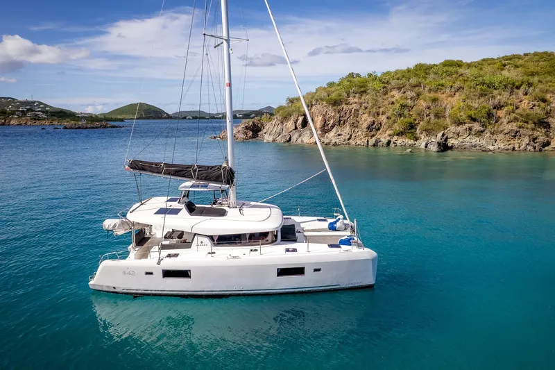 Good Times And Tan Lines Yacht Photos Pics 2023 Lagoon 42 catamaran sailing in clear blue waters near a rocky coastline.