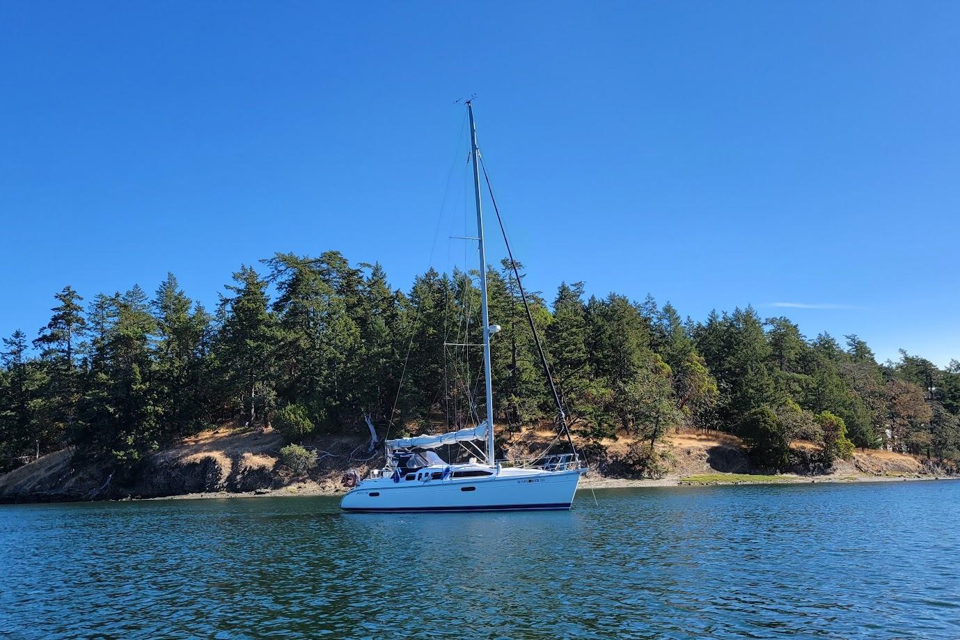 Sailboat Hunter 336 (1995) on calm water near a forested shoreline under clear blue sky.