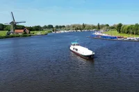 Boat cruising on a river near a windmill and marina, under a clear blue sky.