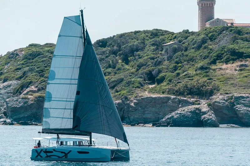  Yacht Photos Pics Manufacturer Provided Image: Sailing catamaran Excess 13, 2026 model, cruising near rocky coastline with lush greenery.