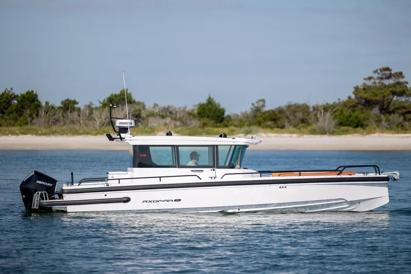  Yacht Photos Pics 2024 Axopar 28 CABIN boat cruising on calm water near a sandy shoreline.