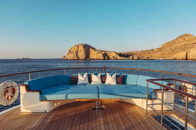 Far Niente Yacht Photos Pics Luxurious 2024 Hoek Design Motor Yacht deck with blue seating, ocean view, and rocky coastline.