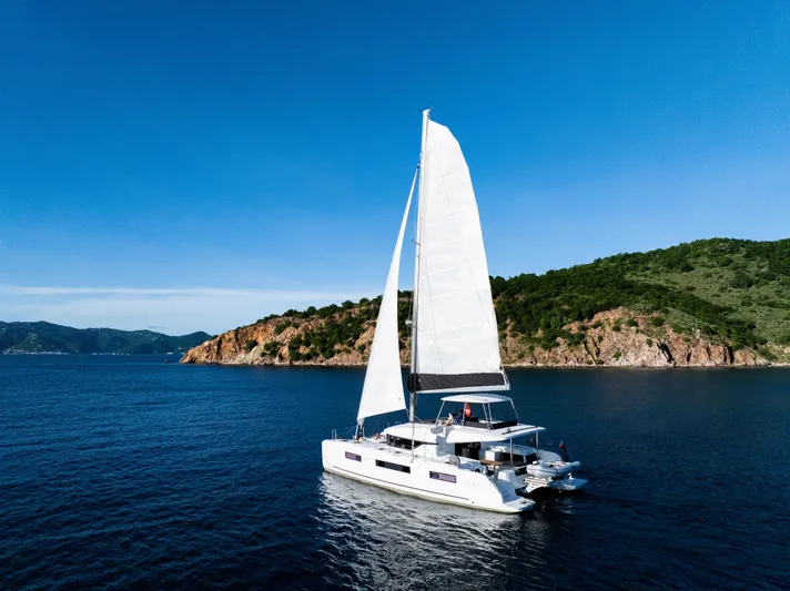Slinky Malinki Yacht Photos Pics 2025 Lagoon 51 catamaran sailing near a rocky coastline under a clear blue sky.