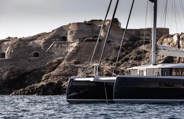  Yacht Photos Pics Catamaran sailing near rocky coastline, Eyecat 55 Autumn 2024 model, under a cloudy sky.