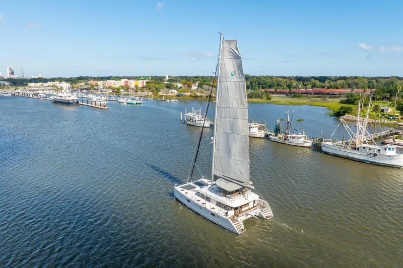 Worth The Wait Yacht Photos Pics Lagoon 560 catamaran sailing in a scenic harbor, 2012 model, surrounded by boats and lush landscape.