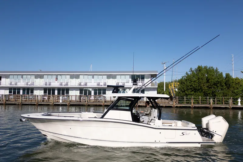 Natural 9 Yacht Photos Pics 2024 Scout 330 LXF boat cruising on water near a modern waterfront building.
