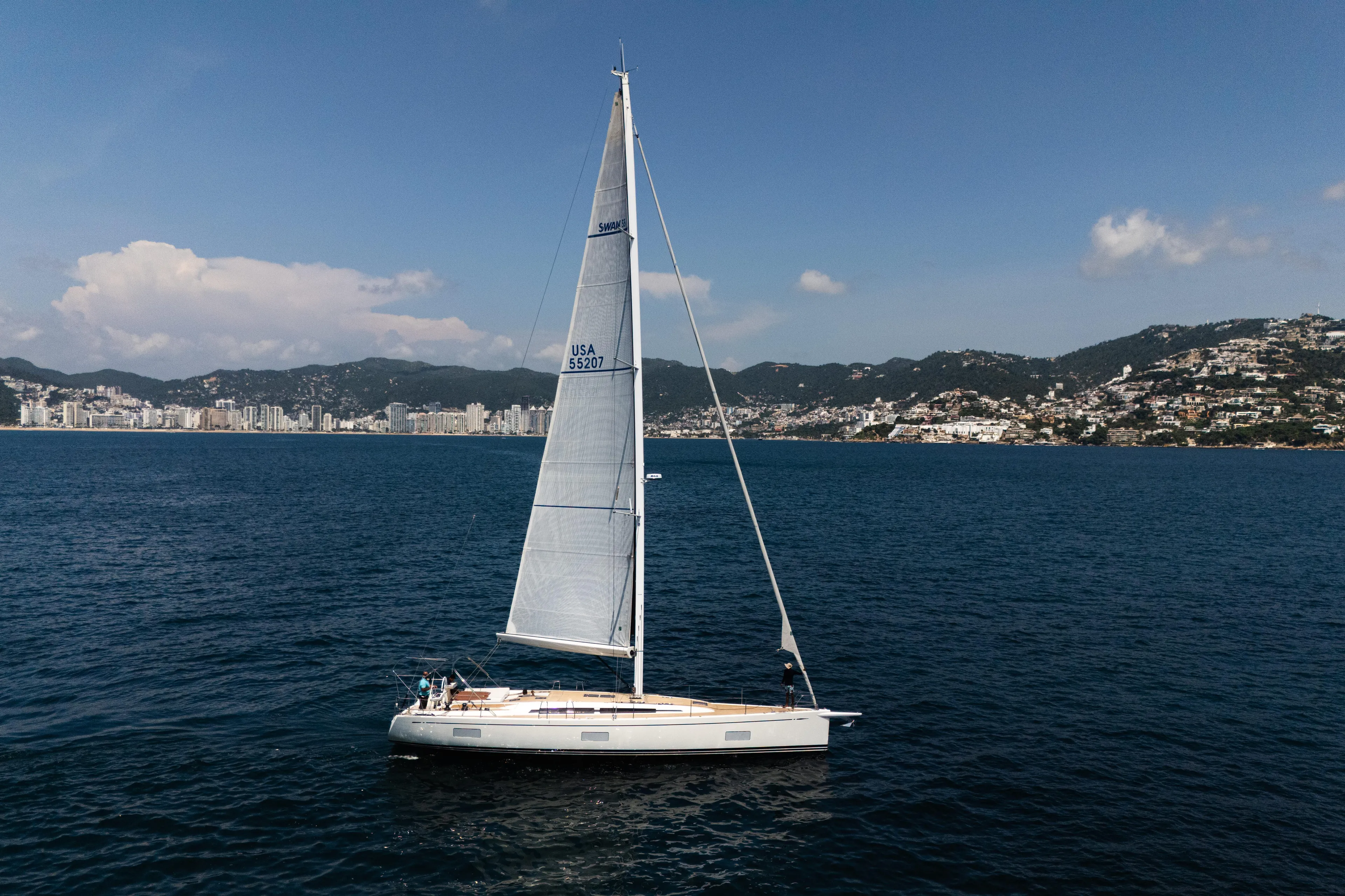 Invictus Yacht Photos Pics 2023 Nautor Swan 55 sailing near coastal cityscape under clear blue sky.