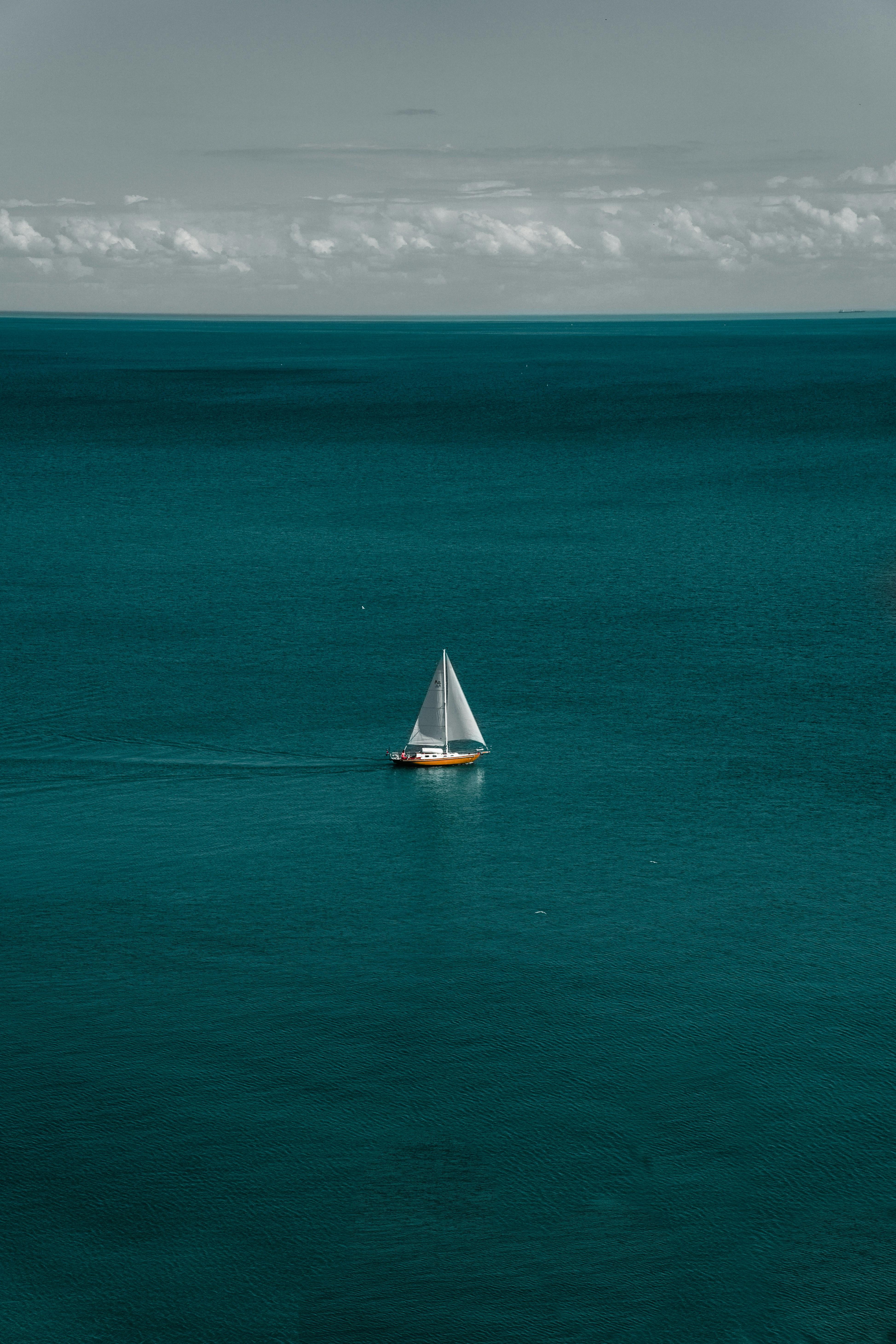 A solitary sailboat on vast, calm turquoise sea under a cloudy sky.