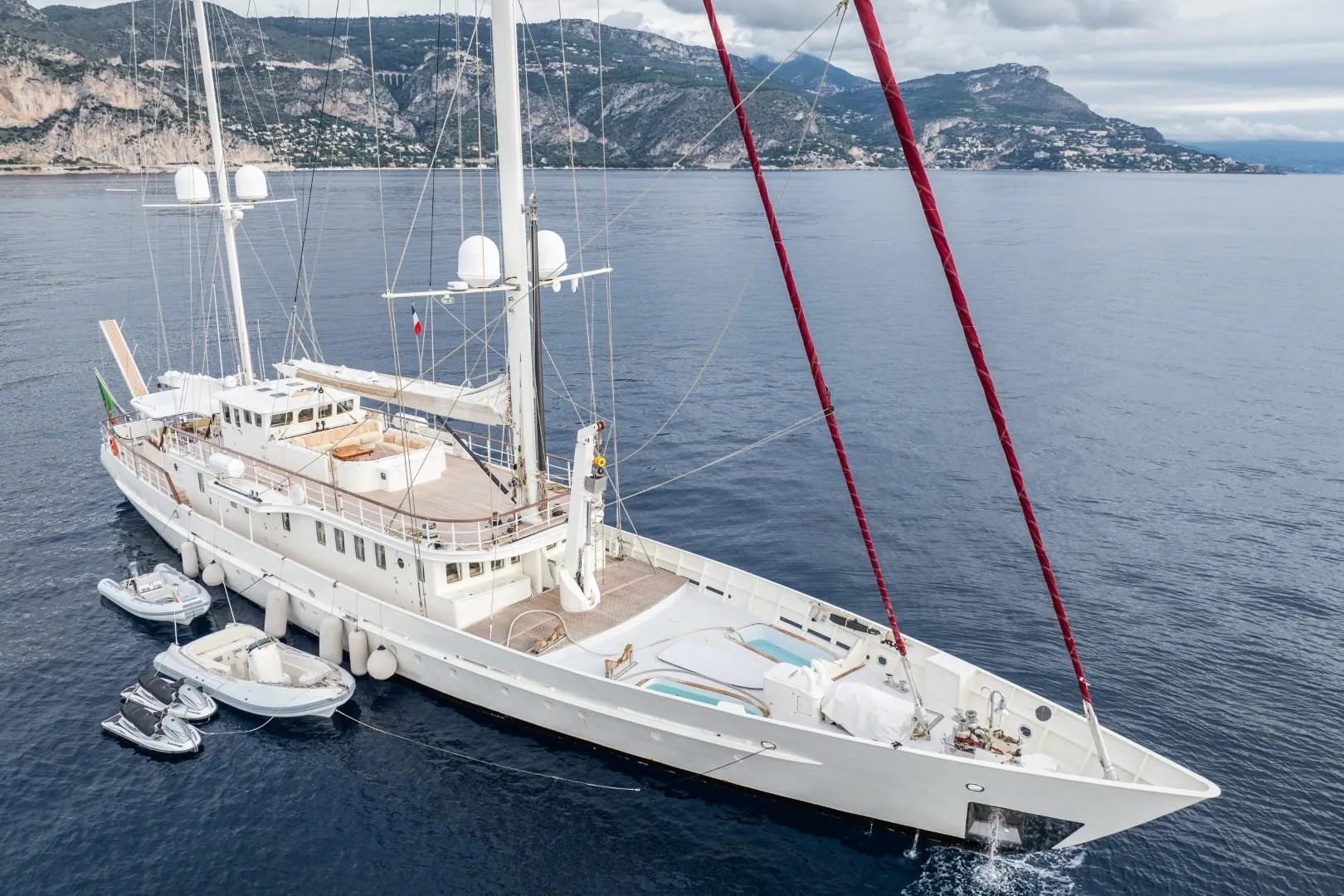 Luxurious custom yacht sailing near scenic coastline, 1990 model, with multiple decks and accompanying boats.