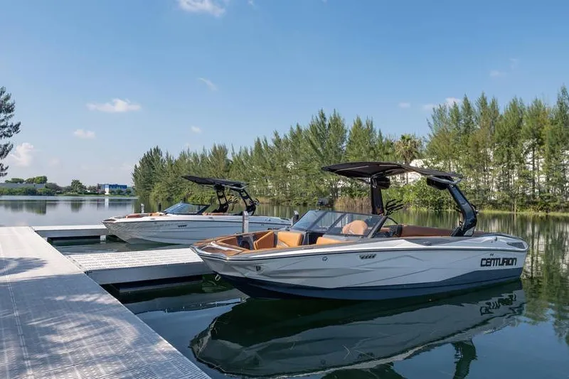  Yacht Photos Pics Two Centurion Nv233 boats docked on a serene lake, surrounded by trees, under a clear sky.