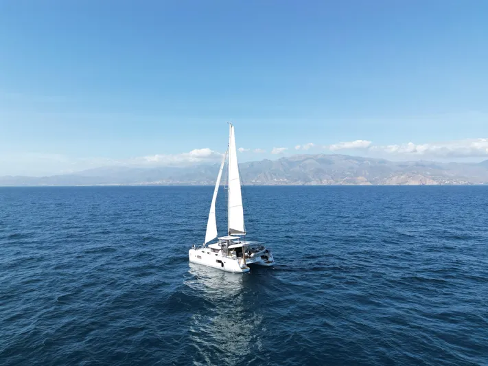  Yacht Photos Pics 2023 Lagoon 42 catamaran sailing on open sea with mountainous coastline in background.