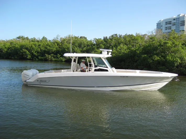 Yacht Photos Pics 2018 Boston Whaler 380 Outrage boat on calm water, surrounded by lush greenery.