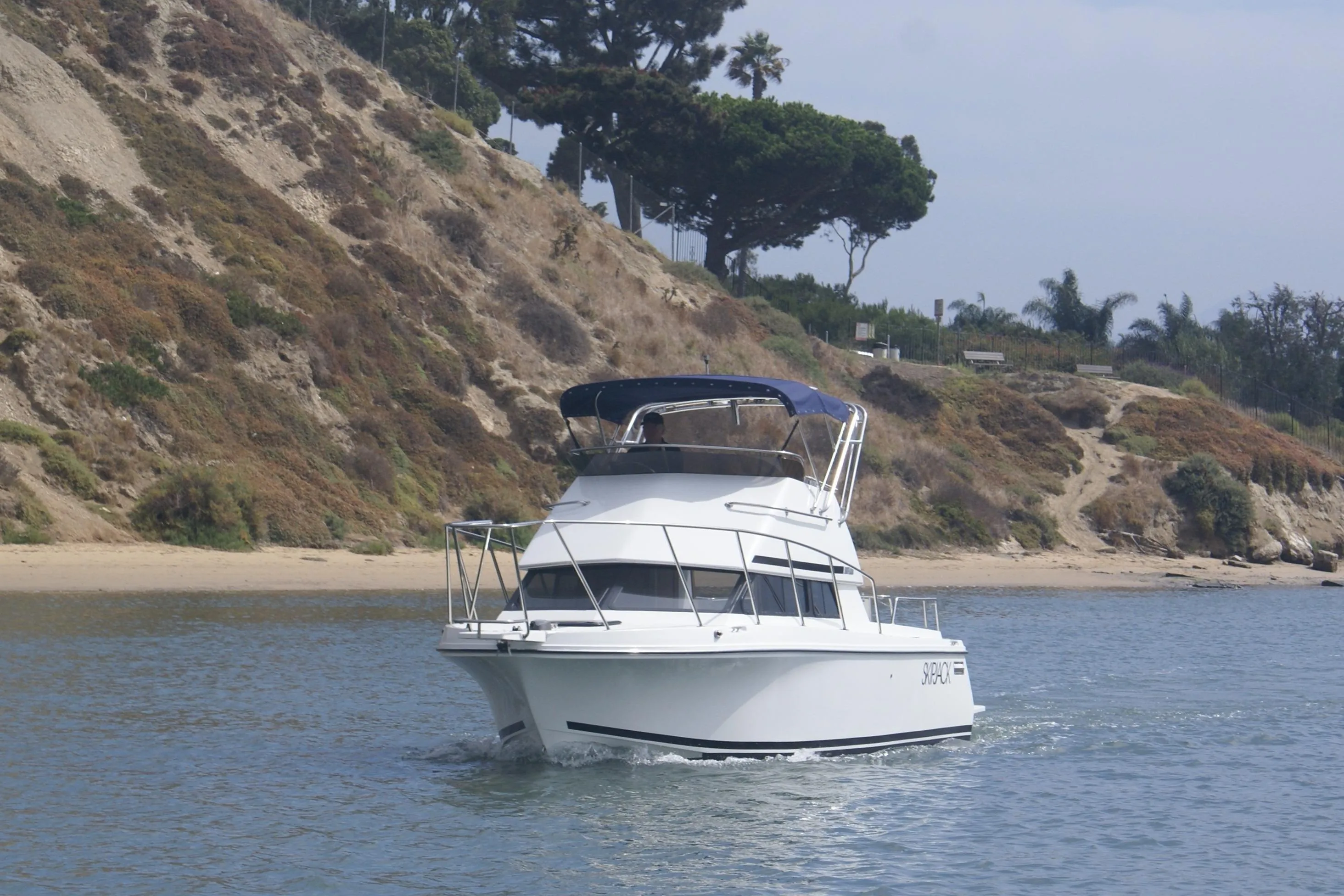 1997 Skipjack 262 Flybridge boat cruising near a rocky shoreline.