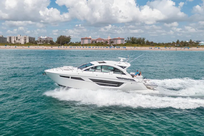 Risky Business Yacht Photos Pics 2019 Cruisers Yachts Cantius sailing near a scenic beach with buildings in the background.