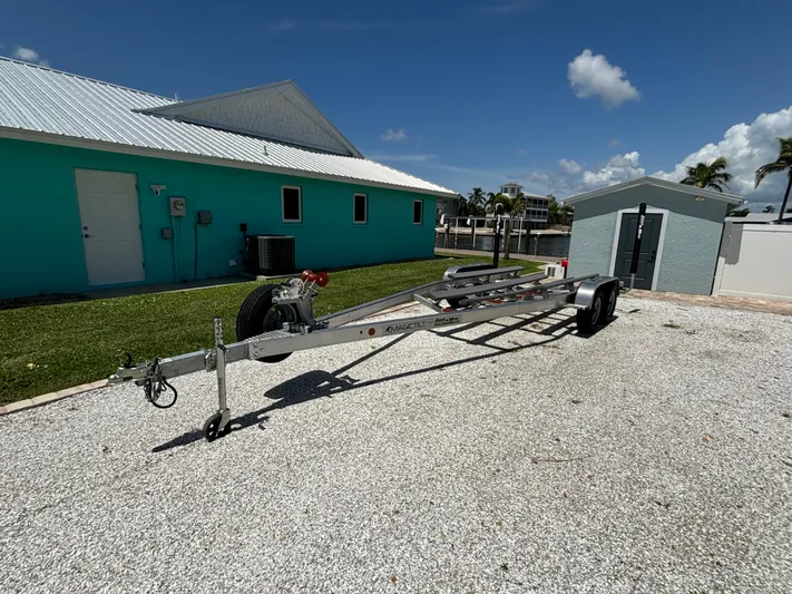  Yacht Photos Pics 2024 Lake & Bay 25 Open boat trailer parked on gravel near turquoise building.