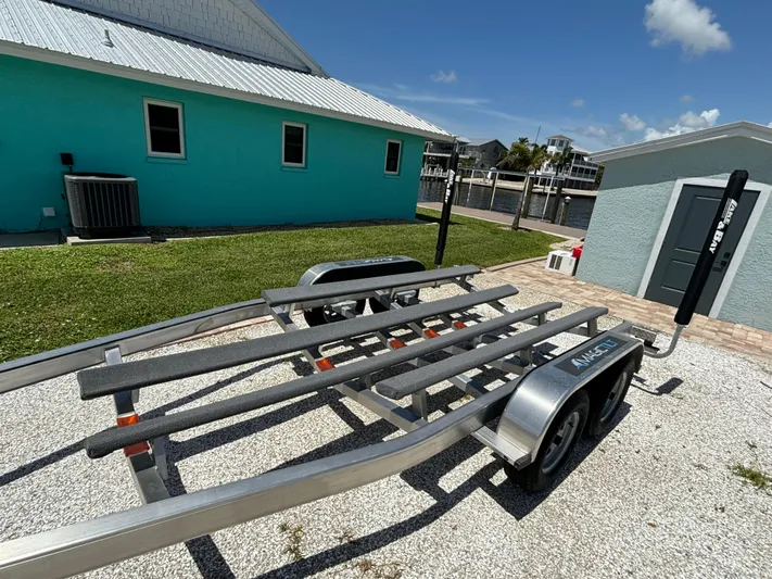  Yacht Photos Pics 2024 Lake & Bay 25 Open boat trailer parked on gravel near turquoise building.