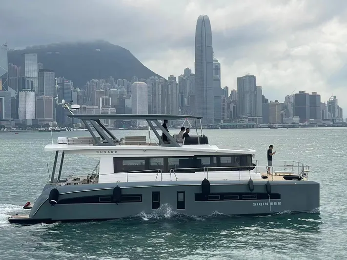  Yacht Photos Pics 2024 Mazarin 55 Catamaran Hybrid cruising in urban harbor with city skyline backdrop.