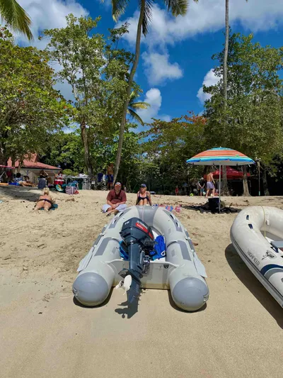 Navicula Yacht Photos Pics Inflatable boat on sandy beach with people relaxing under umbrellas, surrounded by lush trees.