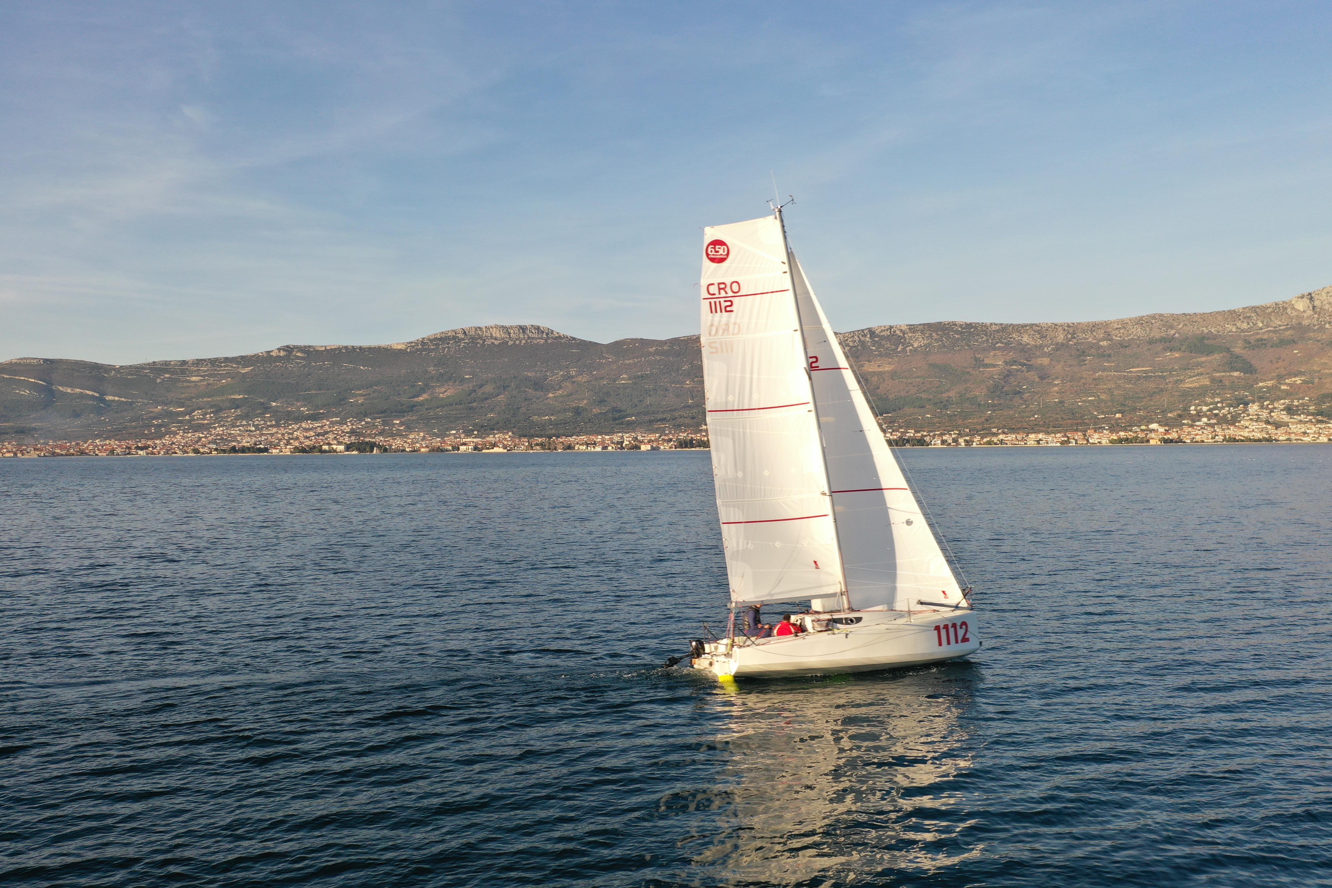 2024 Mini 650 sailboat on calm sea with mountainous coastline in background.
