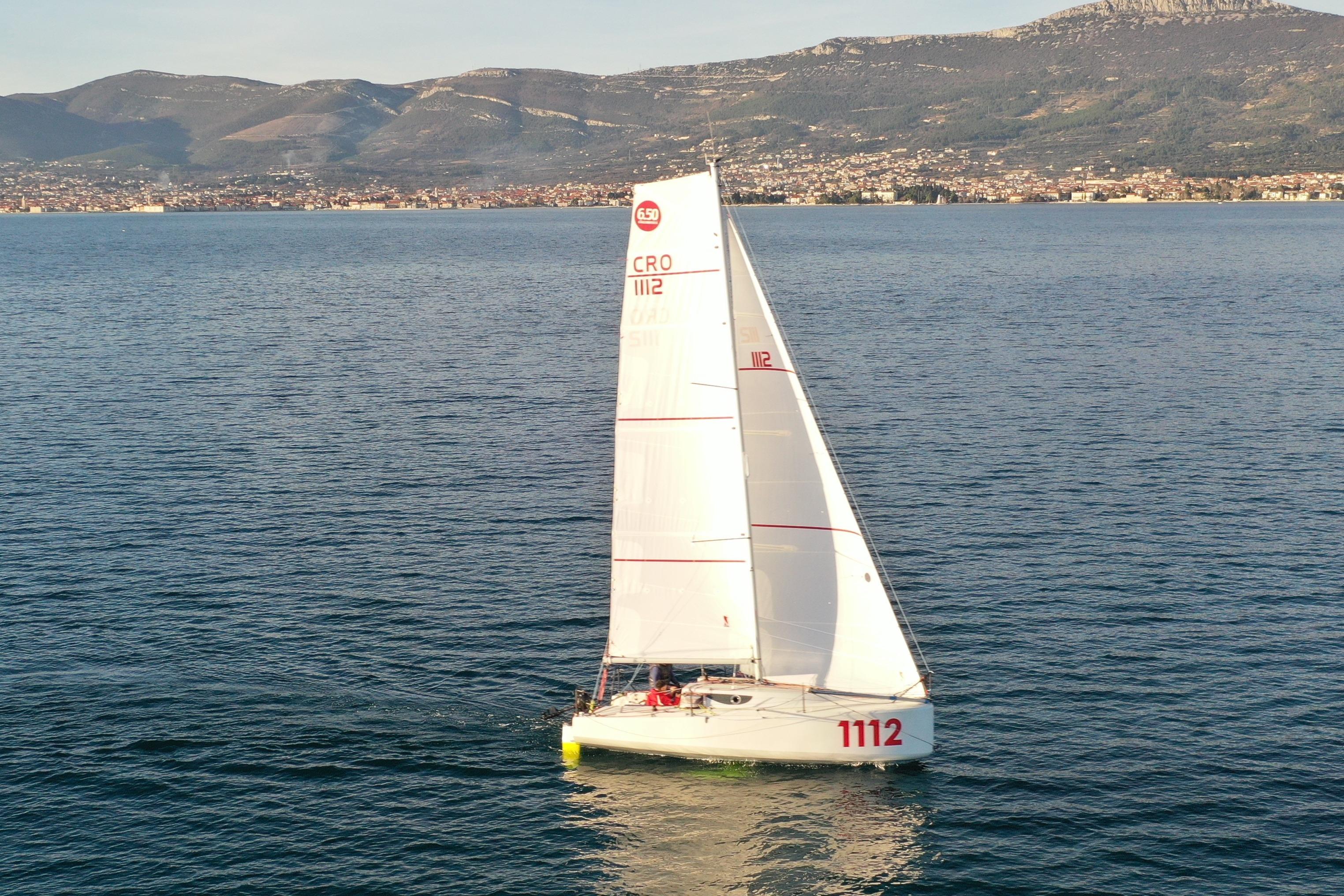 Sailing boat Mini 650, 2024 model, gliding on calm sea with mountainous backdrop.