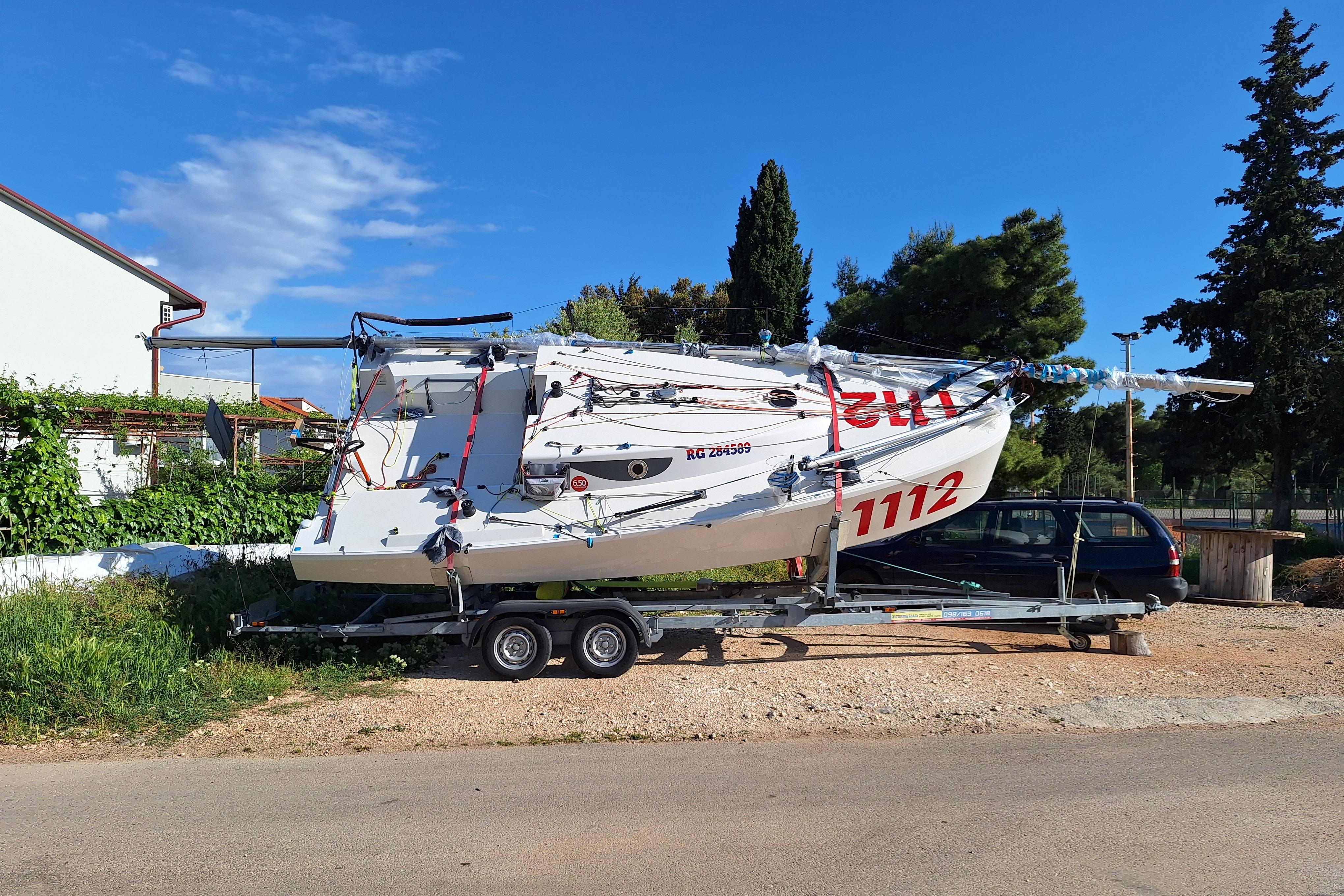 2024 Mini 650 sailboat on trailer, parked outdoors, surrounded by greenery and trees.