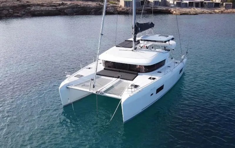  Yacht Photos Pics 2024 Lagoon 42 catamaran sailing in clear blue waters near rocky shoreline.