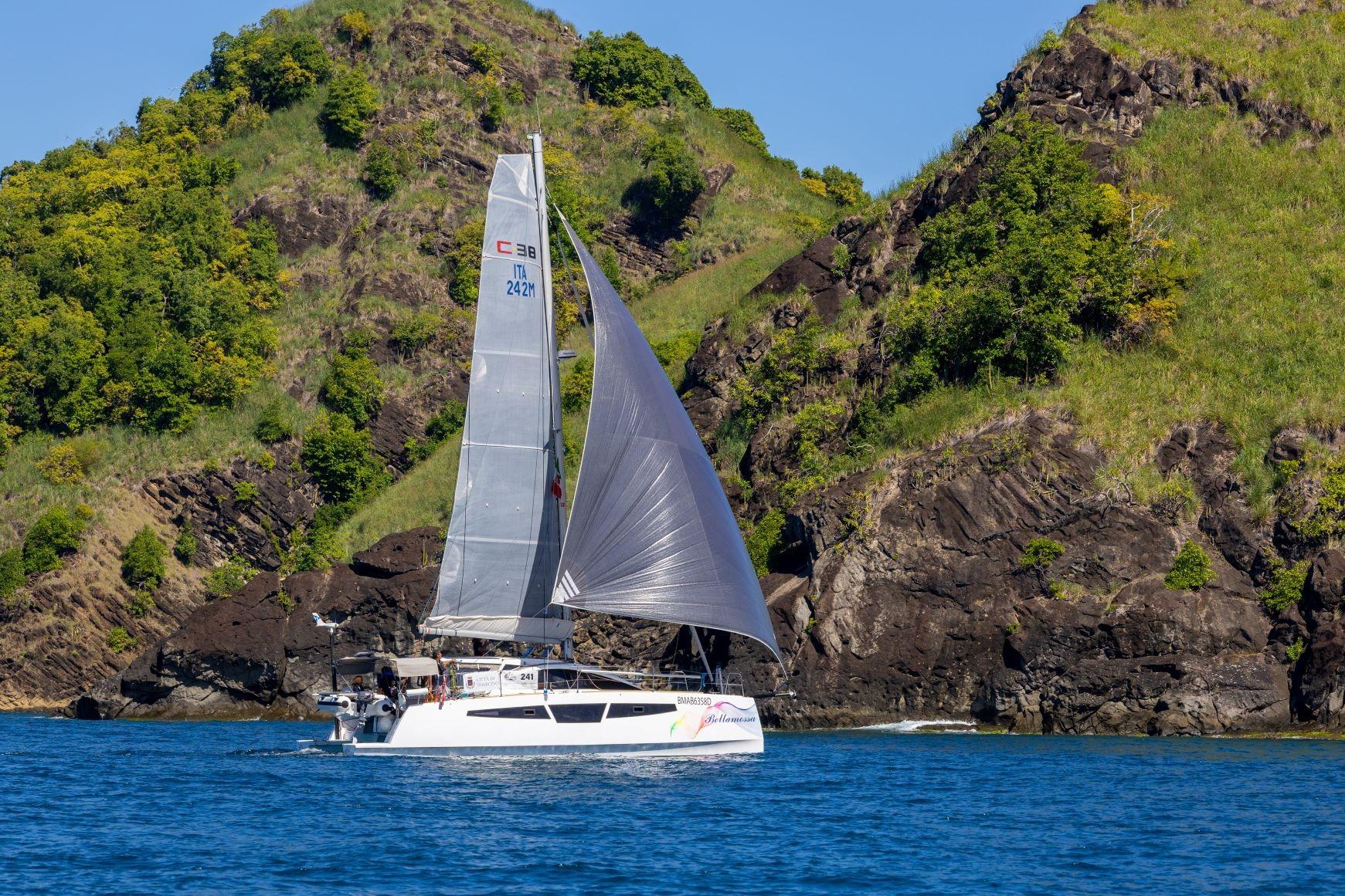 2026 C-Catamarans C-CAT 38 sailing near rocky, green cliffs in clear blue water.