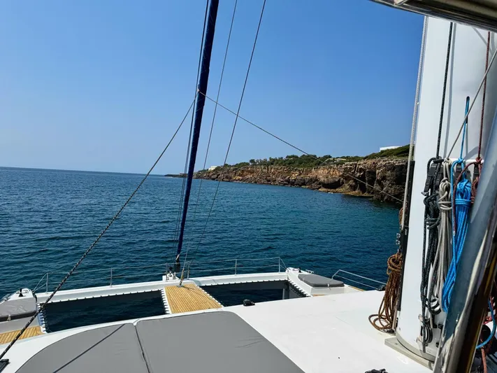 Feel The Blue Yacht Photos Pics Sailing on a 2018 Sunreef 60 catamaran near a rocky coastline under a clear blue sky.