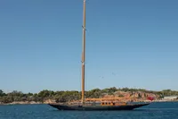 1929 Camper & Nicholsons J Class yacht sailing near a rocky coastline under clear blue skies.