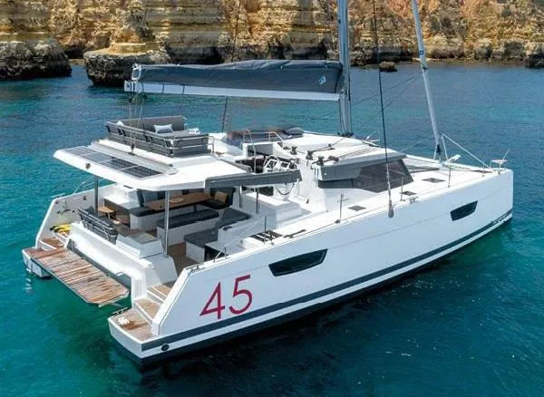  Yacht Photos Pics 2024 Fountaine Pajot Catamaran Elba 45 sailing in clear blue waters near rocky cliffs.