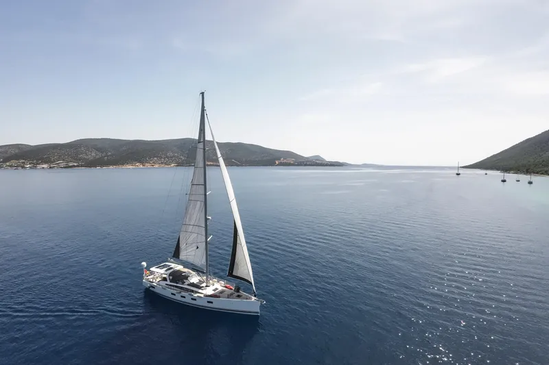  Yacht Photos Pics 2020 Jeanneau 64 sailing yacht on serene blue waters, surrounded by distant hills.